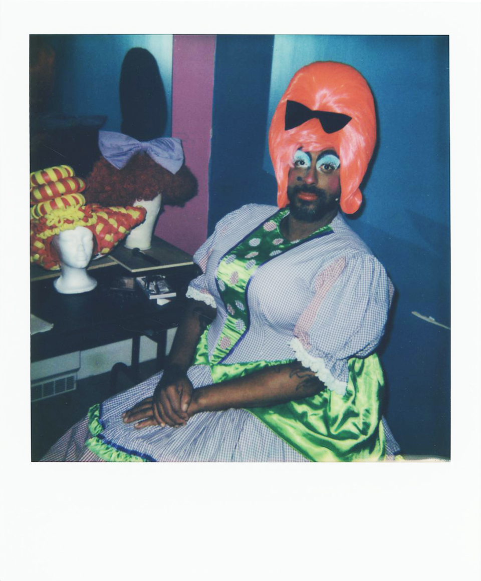 A person in exaggerated drag makeup and a large, bright pink wig with a black bow poses in a colorful dress. Wigs and costume items are visible on a nearby table in the background.